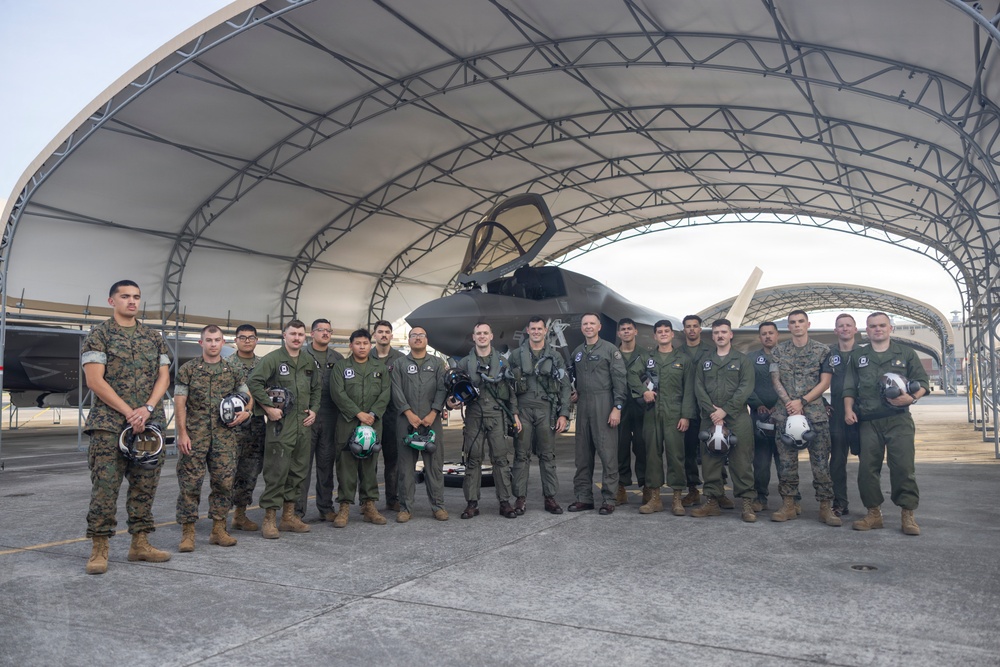 A group of approximately 20 military personnel, appearing to be U.S. Marines and aviators, pose for a group photograph in front of what appears to be an F-35 stealth fighter jet housed under a large arched hangar structure. The personnel are wearing a mix of olive drab flight suits and Marine Corps combat utility uniforms (MARPAT), with several holding flight helmets. The setting appears to be a military air station flight line.