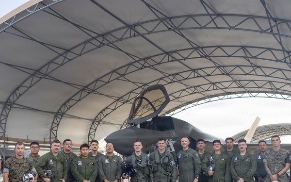 A group of approximately 20 military personnel, appearing to be U.S. Marines and aviators, pose for a group photograph in front of what appears to be an F-35 stealth fighter jet housed under a large arched hangar structure. The personnel are wearing a mix of olive drab flight suits and Marine Corps combat utility uniforms (MARPAT), with several holding flight helmets. The setting appears to be a military air station flight line.