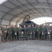 A group of approximately 20 military personnel, appearing to be U.S. Marines and aviators, pose for a group photograph in front of what appears to be an F-35 stealth fighter jet housed under a large arched hangar structure. The personnel are wearing a mix of olive drab flight suits and Marine Corps combat utility uniforms (MARPAT), with several holding flight helmets. The setting appears to be a military air station flight line.