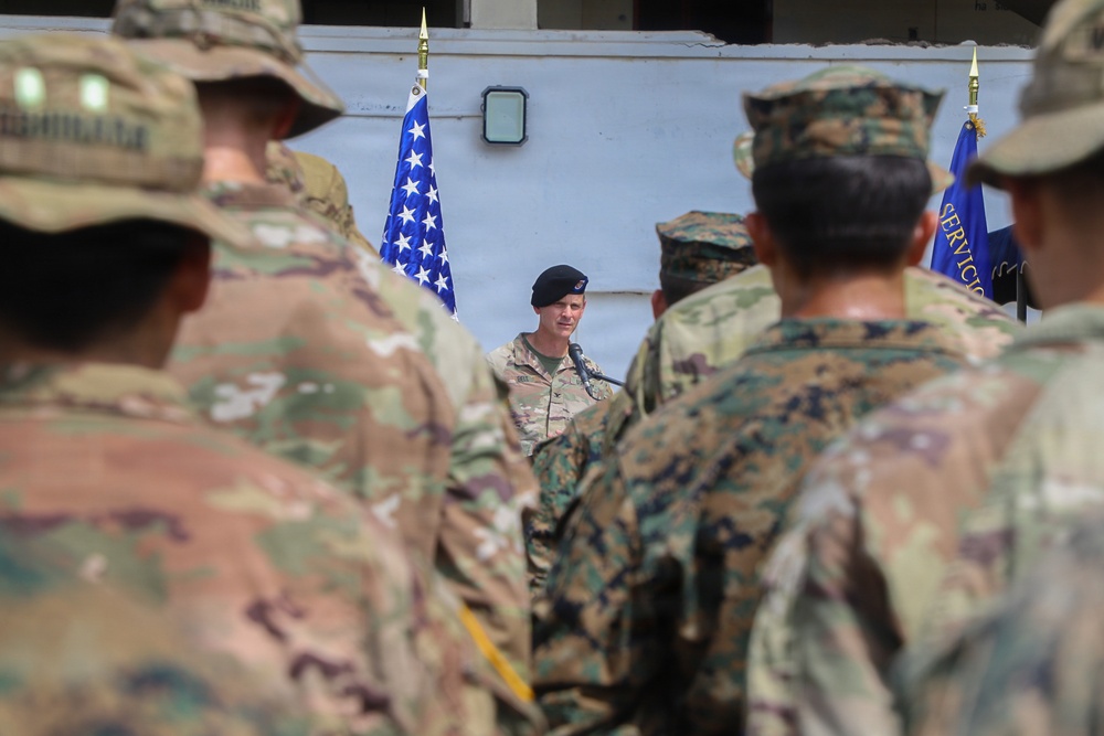 A military ceremony or formation is taking place outdoors, with a senior officer wearing a black beret addressing assembled troops at a microphone. Soldiers in various camouflage uniforms, including OCP (Army) and MARPAT (Marine Corps) patterns, are gathered facing the speaker. An American flag and what appears to be a unit guidon are visible in the background.
