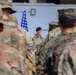 A military ceremony or formation is taking place outdoors, with a senior officer wearing a black beret addressing assembled troops at a microphone. Soldiers in various camouflage uniforms, including OCP (Army) and MARPAT (Marine Corps) patterns, are gathered facing the speaker. An American flag and what appears to be a unit guidon are visible in the background.