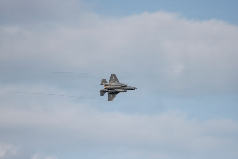 A single stealth fighter jet, consistent with an F-35 Lightning II, is captured in flight against a partly cloudy sky. The aircraft appears to be in a banking or rolling maneuver, viewed from below at a slight angle. Thin contrail or vapor lines are faintly visible extending horizontally across the frame near the aircraft.
