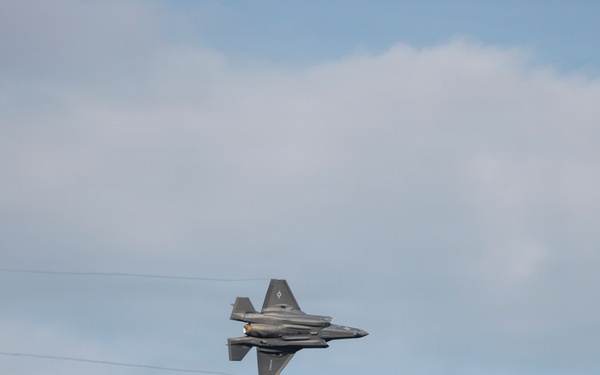 A single stealth fighter jet, consistent with an F-35 Lightning II, is captured in flight against a partly cloudy sky. The aircraft appears to be in a banking or rolling maneuver, viewed from below at a slight angle. Thin contrail or vapor lines are faintly visible extending horizontally across the frame near the aircraft.