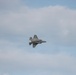 A single stealth fighter jet, consistent with an F-35 Lightning II, is captured in flight against a partly cloudy sky. The aircraft appears to be in a banking or rolling maneuver, viewed from below at a slight angle. Thin contrail or vapor lines are faintly visible extending horizontally across the frame near the aircraft.