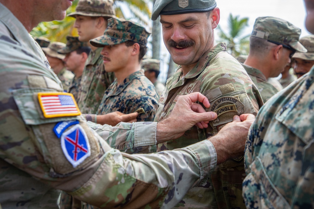 A group of military personnel from multiple branches are gathered outdoors in a tropical setting, appearing to participate in a patch or badge pinning ceremony. A smiling soldier wearing a green beret and OCP (Operational Camouflage Pattern) uniform is receiving what appears to be a 'Jungle Warfare Specialist' tab or patch on his sleeve. Personnel from at least two branches are visible, including U.S. Army and U.S. Marine Corps.