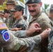A group of military personnel from multiple branches are gathered outdoors in a tropical setting, appearing to participate in a patch or badge pinning ceremony. A smiling soldier wearing a green beret and OCP (Operational Camouflage Pattern) uniform is receiving what appears to be a 'Jungle Warfare Specialist' tab or patch on his sleeve. Personnel from at least two branches are visible, including U.S. Army and U.S. Marine Corps.