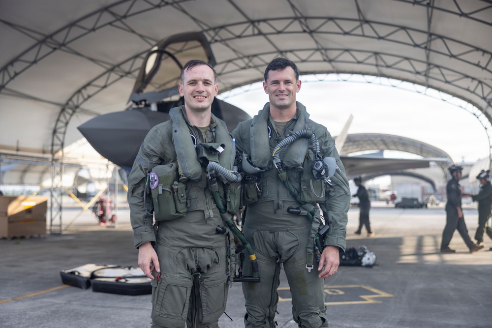 Two military pilots in full flight gear pose for a photo in front of what appears to be an F-35 stealth fighter jet housed under a large hangar canopy. Both are wearing olive green flight suits with survival vests, oxygen mask hoses, and tactical equipment. Additional ground crew personnel are visible in the background.