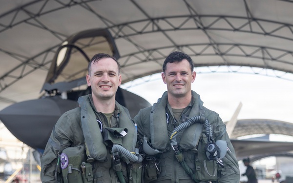 Two military pilots in full flight gear pose for a photo in front of what appears to be an F-35 stealth fighter jet housed under a large hangar canopy. Both are wearing olive green flight suits with survival vests, oxygen mask hoses, and tactical equipment. Additional ground crew personnel are visible in the background.