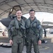 Two military pilots in full flight gear pose for a photo in front of what appears to be an F-35 stealth fighter jet housed under a large hangar canopy. Both are wearing olive green flight suits with survival vests, oxygen mask hoses, and tactical equipment. Additional ground crew personnel are visible in the background.