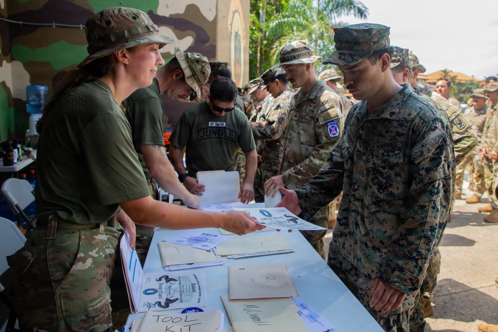 Military personnel from multiple branches are gathered at an outdoor processing or administrative table. A female soldier in multicam uniform is distributing documents to personnel in line, including a U.S. Marine in MARPAT uniform and soldiers wearing 10th Mountain Division patches. The setting appears to be a tropical or subtropical location based on the palm trees and building murals visible in the background.