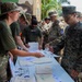 Military personnel from multiple branches are gathered at an outdoor processing or administrative table. A female soldier in multicam uniform is distributing documents to personnel in line, including a U.S. Marine in MARPAT uniform and soldiers wearing 10th Mountain Division patches. The setting appears to be a tropical or subtropical location based on the palm trees and building murals visible in the background.