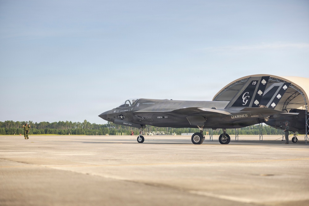 A U.S. Marines F-35B stealth fighter jet is parked on a tarmac near a hangar structure. The aircraft belongs to VMFA-231 squadron, featuring distinctive special livery with spade card suit markings, 'CG' initials, and the number 19 on the tail. Ground crew personnel are visible near the aircraft in what appears to be a ceremonial or commemorative display setting.
