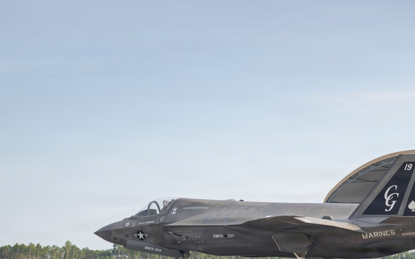 A U.S. Marines F-35B stealth fighter jet is parked on a tarmac near a hangar structure. The aircraft belongs to VMFA-231 squadron, featuring distinctive special livery with spade card suit markings, 'CG' initials, and the number 19 on the tail. Ground crew personnel are visible near the aircraft in what appears to be a ceremonial or commemorative display setting.