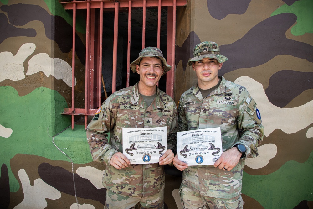 Two U.S. Army soldiers are posing proudly in front of a camouflage-painted wall, each holding a diploma from the Combined Jungle Training Center - South. Both soldiers are wearing OCP (Operational Camouflage Pattern) uniforms with jungle/boonie hats and have earned 'Jungle Expert' certifications from the Jungle Operations Training Course. Their name tapes read 'BONILLA' (left) and 'VALENTIN' (right).