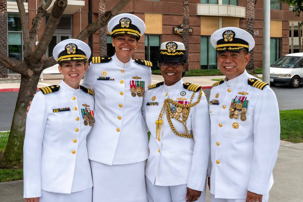 Four U.S. Navy officers in white dress uniforms pose together outdoors, smiling for a group photo. They display various medals, ribbons, and rank insignia consistent with senior officer grades. The setting appears to be near a military or government facility.