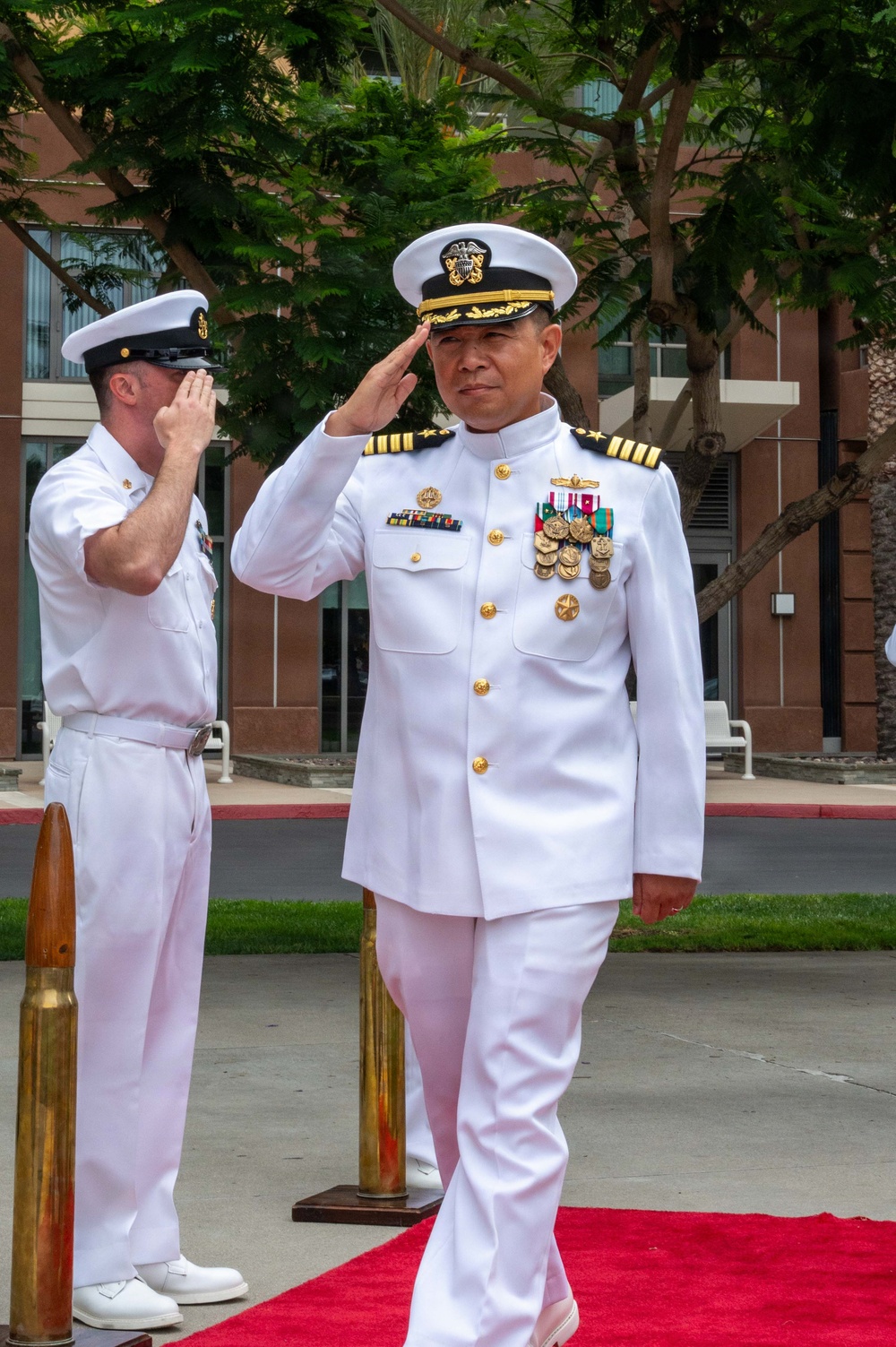 A senior U.S. Navy officer in white dress uniform is rendering a salute while standing on a red carpet during what appears to be a formal military ceremony. The officer displays extensive medal and ribbon decorations consistent with a high-ranking position, with a second Navy enlisted sailor saluting in the background. Large brass artillery shell casings are visible as ceremonial props flanking the red carpet.