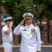 A senior U.S. Navy officer in white dress uniform is rendering a salute while standing on a red carpet during what appears to be a formal military ceremony. The officer displays extensive medal and ribbon decorations consistent with a high-ranking position, with a second Navy enlisted sailor saluting in the background. Large brass artillery shell casings are visible as ceremonial props flanking the red carpet.