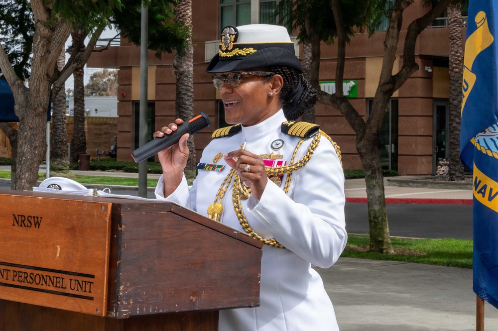 A high-ranking U.S. Navy officer in dress whites is speaking at a podium during an outdoor ceremony. The officer wears a combination cover with gold insignia, shoulder boards indicating senior officer rank (Captain or Admiral), gold aiguillettes, and multiple service ribbons. The podium bears the label 'NRSW' and 'NT PERSONNEL UNIT', suggesting a Naval Reserve or Naval Station Southwest event.