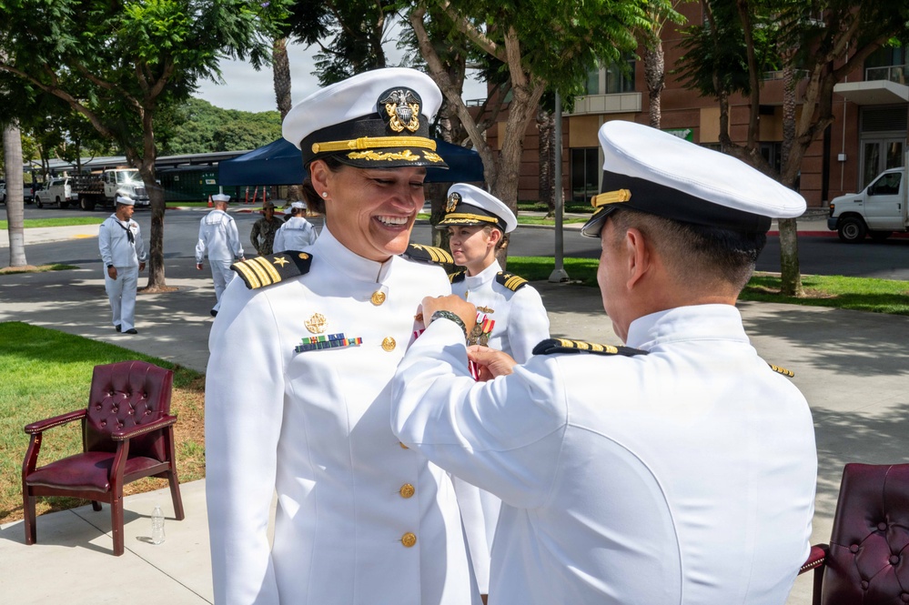 A U.S. Navy military ceremony is taking place outdoors, likely on a naval base. A senior female officer is being presented with a medal or decoration by another officer, both wearing white dress uniforms. Additional naval personnel are visible in the background observing the ceremony.