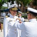 A U.S. Navy military ceremony is taking place outdoors, likely on a naval base. A senior female officer is being presented with a medal or decoration by another officer, both wearing white dress uniforms. Additional naval personnel are visible in the background observing the ceremony.