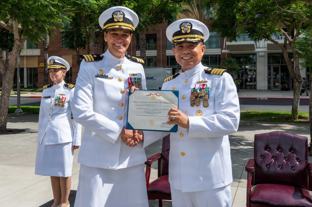 Two senior U.S. Navy officers in white dress uniforms are shaking hands while jointly holding a Meritorious Service Medal certificate from the United States of America. The ceremony appears to be taking place outdoors in an urban setting, likely at a naval installation or government facility. A third naval officer in white dress uniform stands at attention in the background.