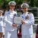 Two senior U.S. Navy officers in white dress uniforms are shaking hands while jointly holding a Meritorious Service Medal certificate from the United States of America. The ceremony appears to be taking place outdoors in an urban setting, likely at a naval installation or government facility. A third naval officer in white dress uniform stands at attention in the background.