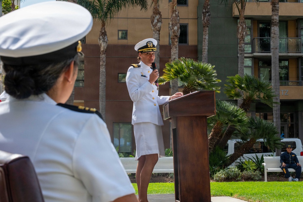 A U.S. Navy officer in white dress uniform is delivering a speech at an outdoor podium, holding a microphone. Another Navy officer in white uniform is seated in the foreground with their back to the camera. The event appears to be a formal military ceremony taking place in an outdoor plaza with palm trees and a multi-story building in the background.