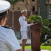A U.S. Navy officer in white dress uniform is delivering a speech at an outdoor podium, holding a microphone. Another Navy officer in white uniform is seated in the foreground with their back to the camera. The event appears to be a formal military ceremony taking place in an outdoor plaza with palm trees and a multi-story building in the background.