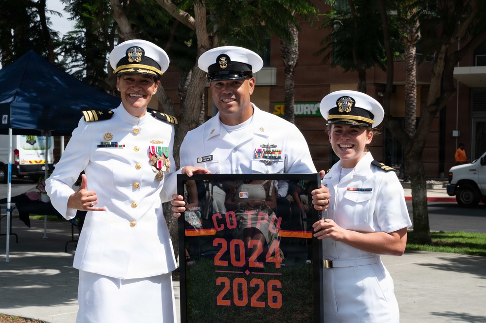 Three U.S. Navy personnel in white dress uniforms pose together outdoors, smiling for a photo. The central figure holds a framed photograph displaying the text 'CO TPU 2024 2026,' suggesting a command or unit milestone. The setting appears to be an outdoor event near a Subway restaurant.