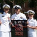 Three U.S. Navy personnel in white dress uniforms pose together outdoors, smiling for a photo. The central figure holds a framed photograph displaying the text 'CO TPU 2024 2026,' suggesting a command or unit milestone. The setting appears to be an outdoor event near a Subway restaurant.