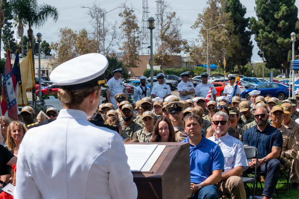 A U.S. Navy officer in dress whites is delivering a speech at a podium during an outdoor military ceremony. The audience consists of a large group of Navy personnel in various uniforms along with civilian attendees seated and standing in an outdoor plaza area. The setting appears to be a naval installation, with a building labeled 'Mercer Hall' visible in the background.