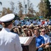 A U.S. Navy officer in dress whites is delivering a speech at a podium during an outdoor military ceremony. The audience consists of a large group of Navy personnel in various uniforms along with civilian attendees seated and standing in an outdoor plaza area. The setting appears to be a naval installation, with a building labeled 'Mercer Hall' visible in the background.