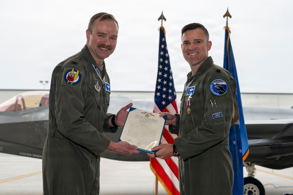 Two U.S. Air Force personnel in olive green flight suits are posing together in an aircraft hangar, holding a formal certificate or commendation document. An F-35 fighter jet is visible in the background along with an American flag and a U.S. Air Force flag. The occasion appears to be a military award or recognition ceremony.