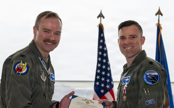 Two U.S. Air Force personnel in olive green flight suits are posing together in an aircraft hangar, holding a formal certificate or commendation document. An F-35 fighter jet is visible in the background along with an American flag and a U.S. Air Force flag. The occasion appears to be a military award or recognition ceremony.