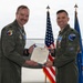 Two U.S. Air Force personnel in olive green flight suits are posing together in an aircraft hangar, holding a formal certificate or commendation document. An F-35 fighter jet is visible in the background along with an American flag and a U.S. Air Force flag. The occasion appears to be a military award or recognition ceremony.