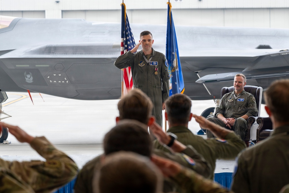 A U.S. Air Force military ceremony, likely a change of command, taking place inside an aircraft hangar. A uniformed officer in olive flight suit stands at attention rendering a salute while assembled personnel in the foreground return the salute. A second officer is seated in a leather chair to the right, smiling, with F-35 stealth fighter jets and American and Air Force flags visible in the background.