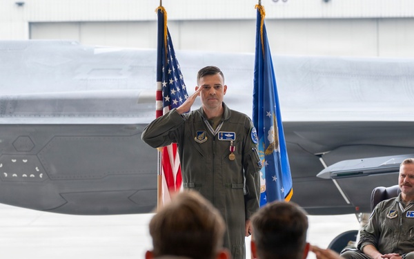A U.S. Air Force military ceremony, likely a change of command, taking place inside an aircraft hangar. A uniformed officer in olive flight suit stands at attention rendering a salute while assembled personnel in the foreground return the salute. A second officer is seated in a leather chair to the right, smiling, with F-35 stealth fighter jets and American and Air Force flags visible in the background.