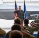 A U.S. Air Force military ceremony, likely a change of command, taking place inside an aircraft hangar. A uniformed officer in olive flight suit stands at attention rendering a salute while assembled personnel in the foreground return the salute. A second officer is seated in a leather chair to the right, smiling, with F-35 stealth fighter jets and American and Air Force flags visible in the background.