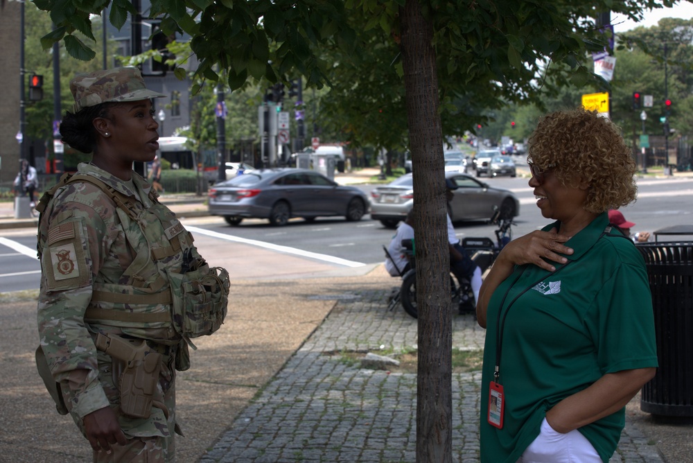 Two women are engaged in conversation on a urban sidewalk. One woman is wearing a U.S. military camouflage uniform (OCP pattern) with a U.S. flag patch and tactical gear, while the other is wearing a green polo shirt with a lanyard/ID badge. The setting appears to be a busy city street with traffic, trees, and traffic signals visible in the background.