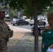 Two women are engaged in conversation on a urban sidewalk. One woman is wearing a U.S. military camouflage uniform (OCP pattern) with a U.S. flag patch and tactical gear, while the other is wearing a green polo shirt with a lanyard/ID badge. The setting appears to be a busy city street with traffic, trees, and traffic signals visible in the background.