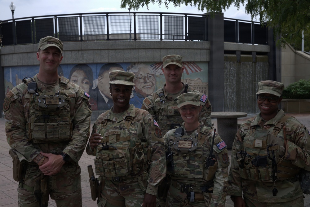 Five U.S. military personnel in OCP (Operational Camouflage Pattern) uniforms pose together in front of a public plaza featuring a colorful mural and a circular fountain structure. The group consists of both male and female service members wearing tactical vests, patrol caps, and American flag patches. The setting appears to be an urban public space, possibly during a deployment or civil support mission.