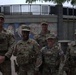 Five U.S. military personnel in OCP (Operational Camouflage Pattern) uniforms pose together in front of a public plaza featuring a colorful mural and a circular fountain structure. The group consists of both male and female service members wearing tactical vests, patrol caps, and American flag patches. The setting appears to be an urban public space, possibly during a deployment or civil support mission.