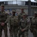 Five military personnel in OCP (Operational Camouflage Pattern) uniforms are posed together in what appears to be an outdoor public plaza or urban area. They are wearing tactical vests and equipment, suggesting they are on an active duty assignment or patrol. A colorful mural depicting civilian figures is visible in the background.