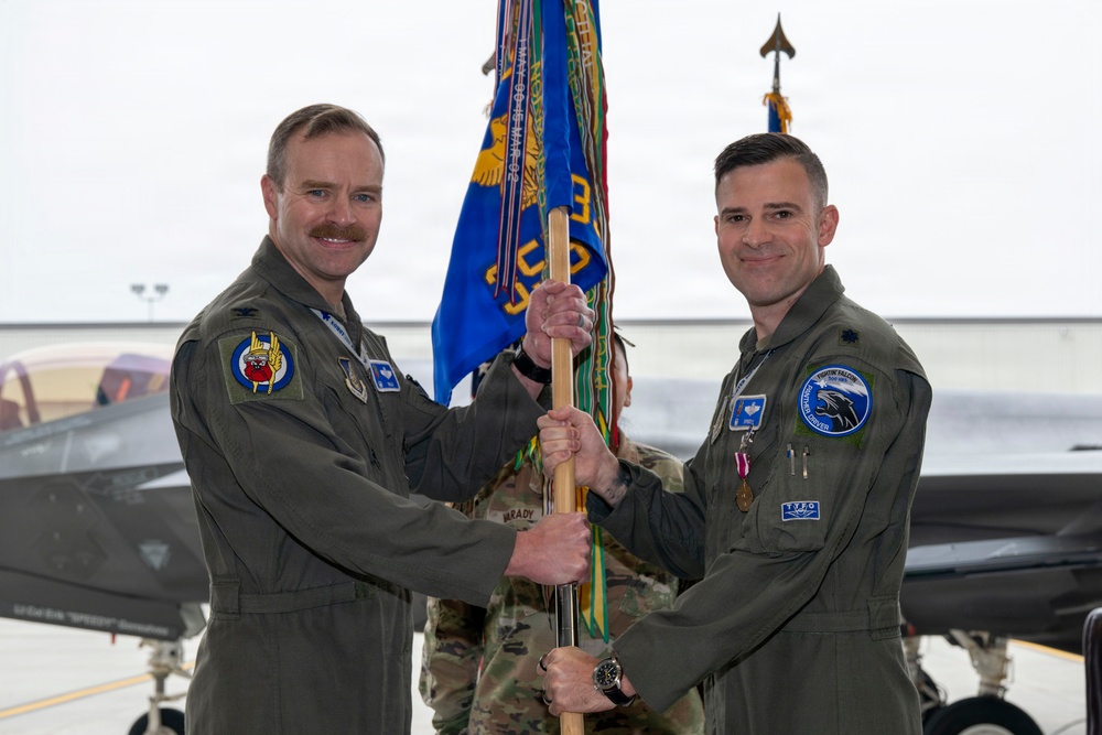 Two U.S. Air Force officers in olive drab flight suits are participating in a military change of command ceremony, jointly holding a unit guidon (flag) in what appears to be an aircraft hangar. The ceremony is taking place in front of what appears to be an F-35 fighter jet. Both officers are wearing rank insignia and unit patches consistent with a formal military ceremony.