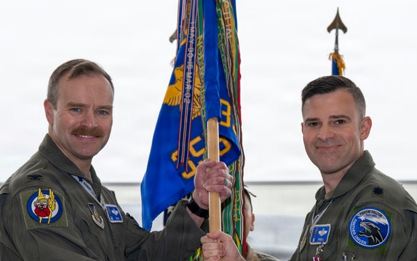 Two U.S. Air Force officers in olive drab flight suits are participating in a military change of command ceremony, jointly holding a unit guidon (flag) in what appears to be an aircraft hangar. The ceremony is taking place in front of what appears to be an F-35 fighter jet. Both officers are wearing rank insignia and unit patches consistent with a formal military ceremony.