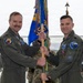 Two U.S. Air Force officers in olive drab flight suits are participating in a military change of command ceremony, jointly holding a unit guidon (flag) in what appears to be an aircraft hangar. The ceremony is taking place in front of what appears to be an F-35 fighter jet. Both officers are wearing rank insignia and unit patches consistent with a formal military ceremony.