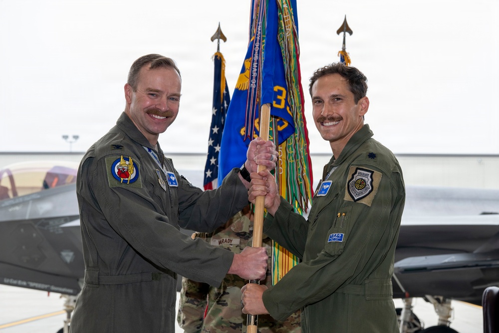 Two U.S. Air Force officers in olive green flight suits are participating in a military guidon/flag transfer ceremony, likely a change of command. They are jointly holding a wooden staff with a unit guidon in front of military flags inside an aircraft hangar. An F-35 fighter jet is visible in the background.