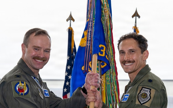 Two U.S. Air Force officers in olive green flight suits are participating in a military guidon/flag transfer ceremony, likely a change of command. They are jointly holding a wooden staff with a unit guidon in front of military flags inside an aircraft hangar. An F-35 fighter jet is visible in the background.