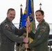 Two U.S. Air Force officers in olive green flight suits are participating in a military guidon/flag transfer ceremony, likely a change of command. They are jointly holding a wooden staff with a unit guidon in front of military flags inside an aircraft hangar. An F-35 fighter jet is visible in the background.