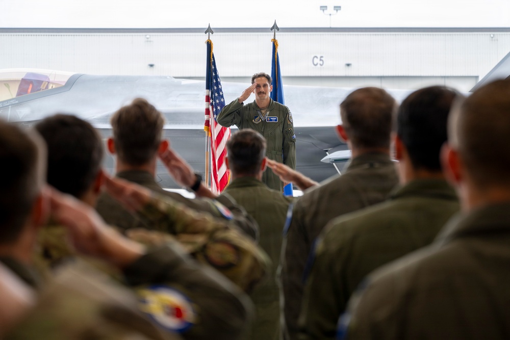 A military officer in a flight suit stands at the front of a hangar, returning a salute to a formation of assembled personnel. The ceremony appears to be a formal military event such as a change of command or unit activation, taking place in an aircraft hangar with a fighter jet visible in the background. Both the American flag and what appears to be a U.S. Air Force flag are prominently displayed behind the officer.